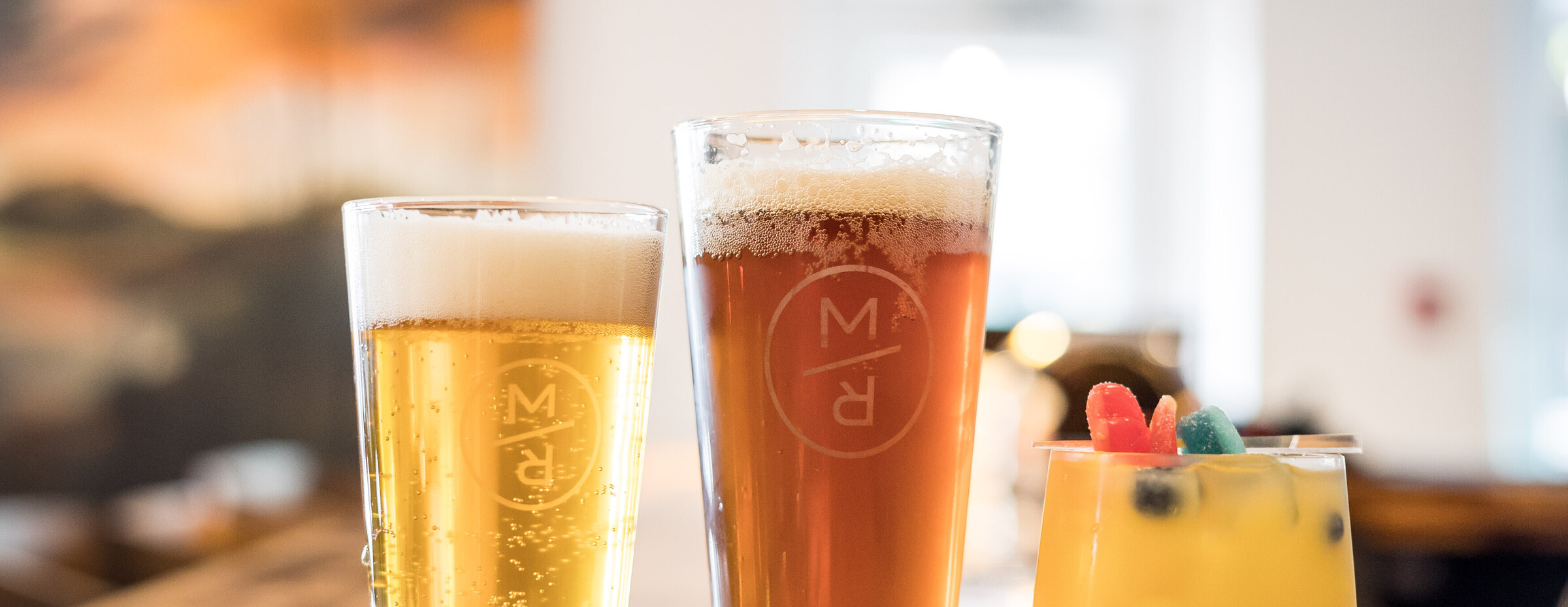 Vus de près, déposés sur un bar, deux verres remplis de bière (arborant le logo de la Microbrasserie Maître Renard de Chibougamau) et un verre à cocktail, étant lui aussi bien rempli.