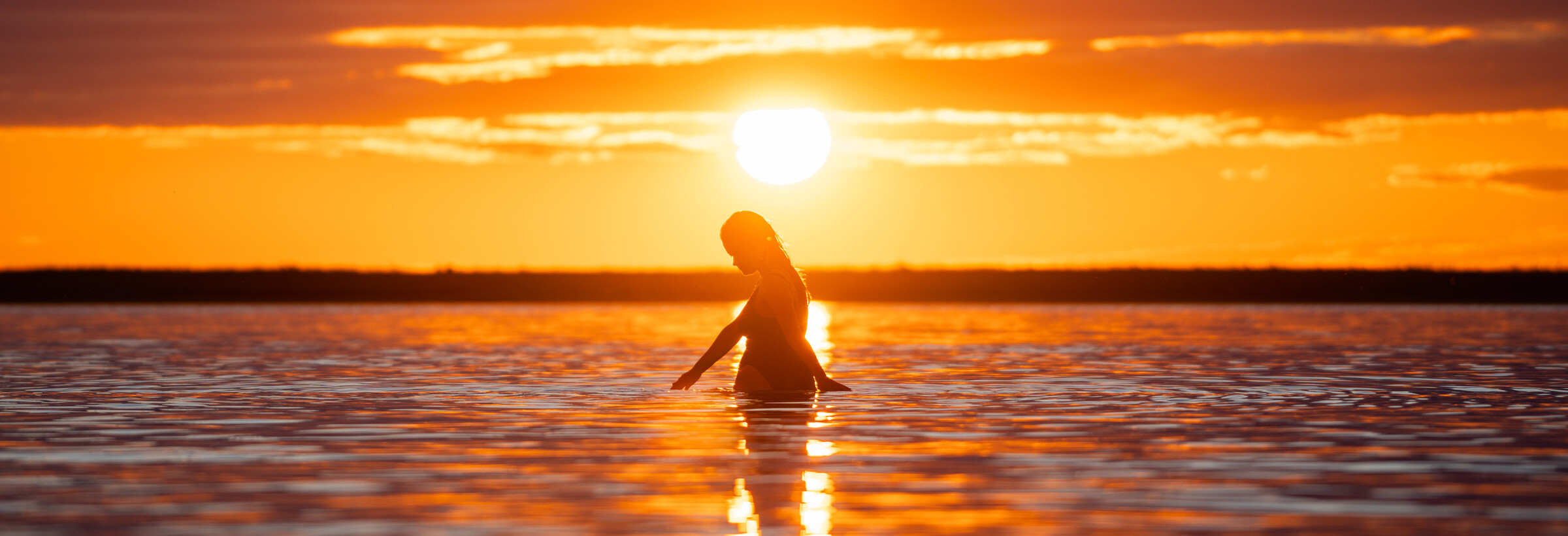Coucher de soleil sur la Baie James. À contre-jour, une femme dans l'eau jusqu'au niveau de la taille, s'amuse en touchant la surface.